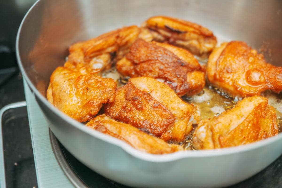 Potongan paha ayam ala Provence berwarna keemasan sedang dimasak dalam wajan dengan minyak panas di atas kompor.