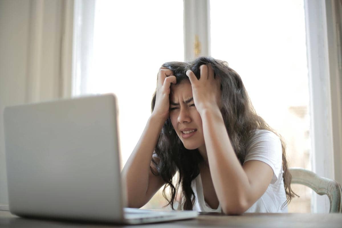 Seorang wanita memegang kepala dengan ekspresi lelah di depan laptop di meja kerja dekat jendela dengan cahaya alami.
