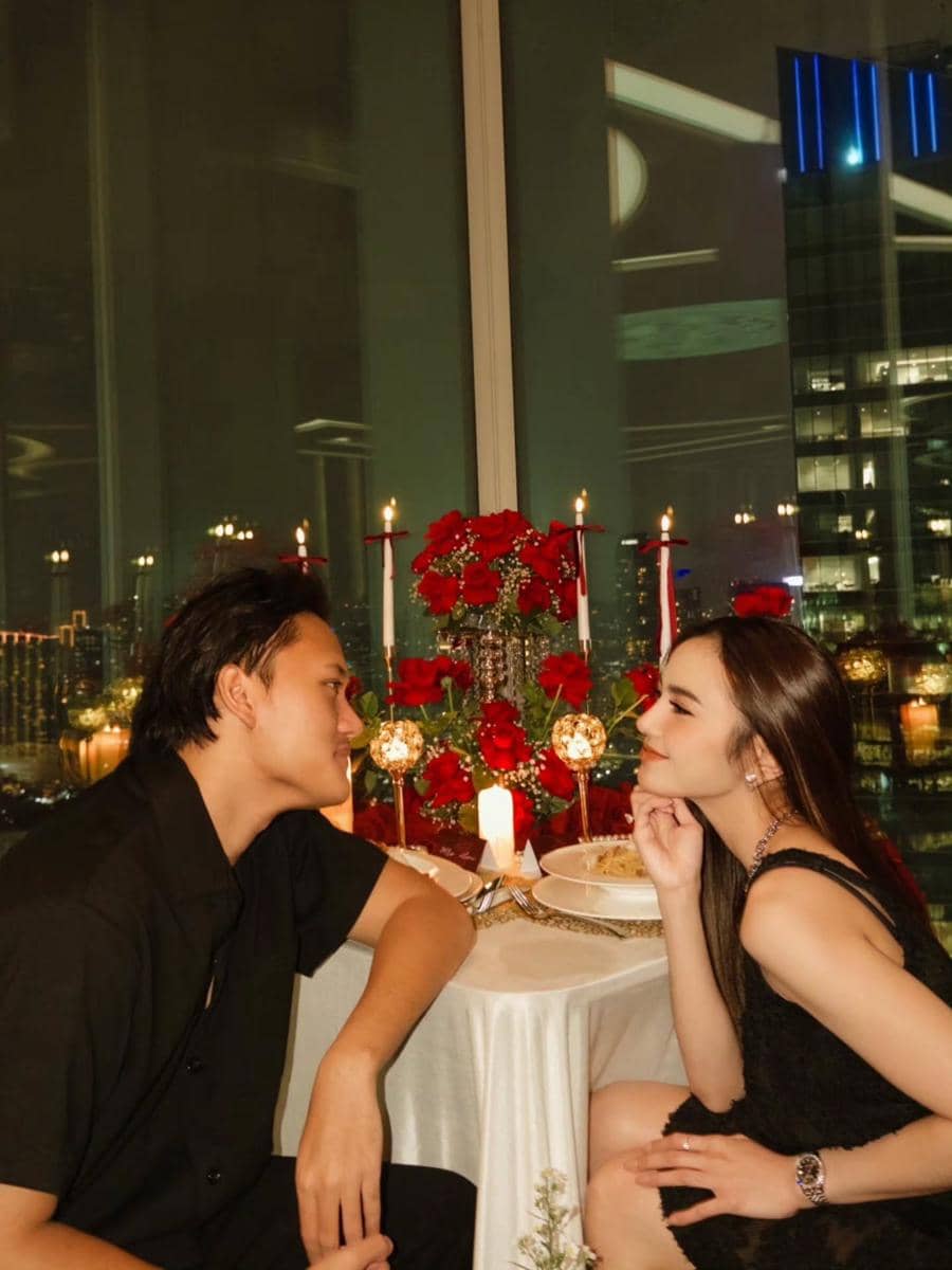 Sepasang kekasih duduk berhadapan di meja makan romantis dengan dekorasi bunga mawar merah dan lilin di restoran berlatarkan gedung kota malam hari.