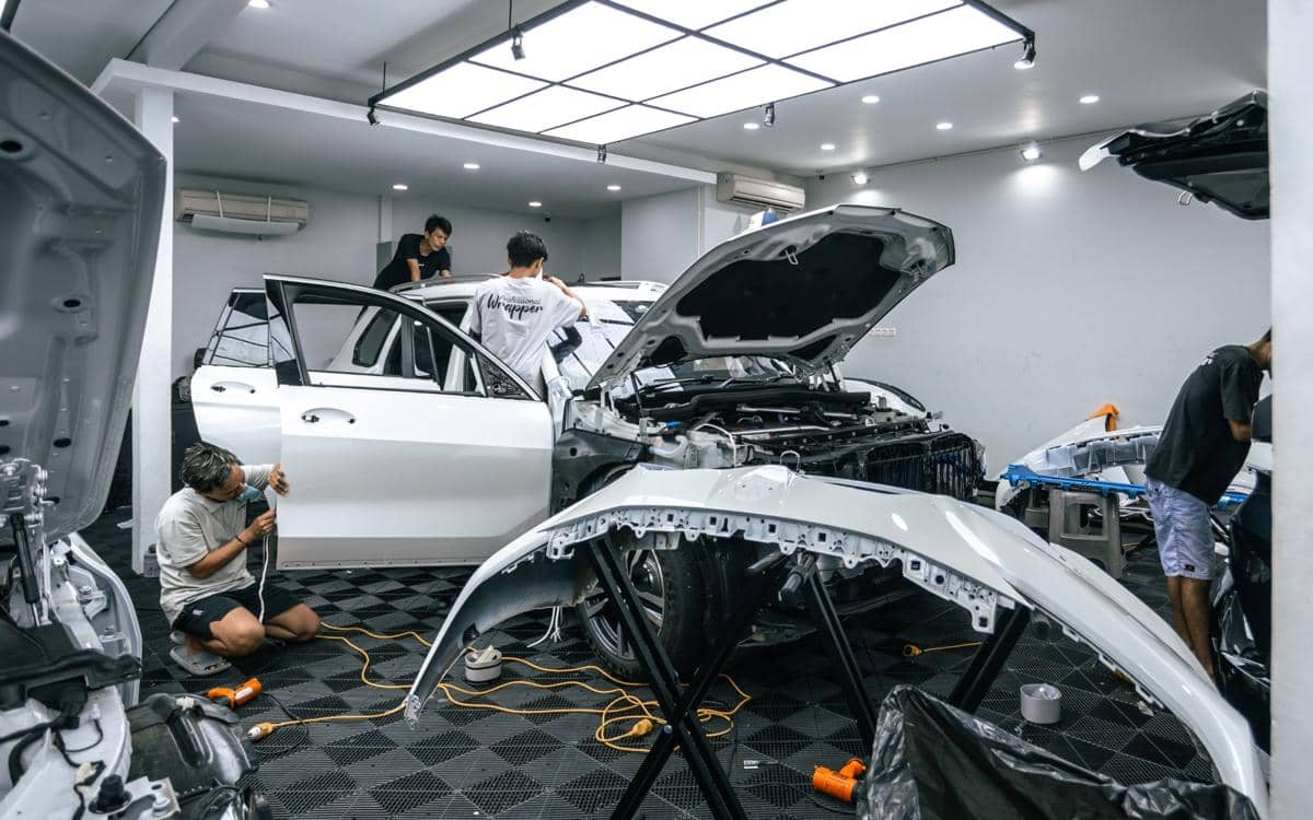 Beberapa teknisi sedang memasang paint protection film (PPF) pada mobil putih di bengkel dengan peralatan lengkap dan pencahayaan terang.