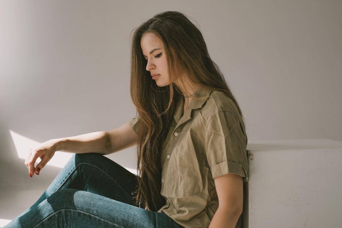 Perempuan berambut panjang duduk dengan ekspresi sedih di ruangan bercahaya lembut, mengenakan kemeja cokelat muda dan celana jeans.