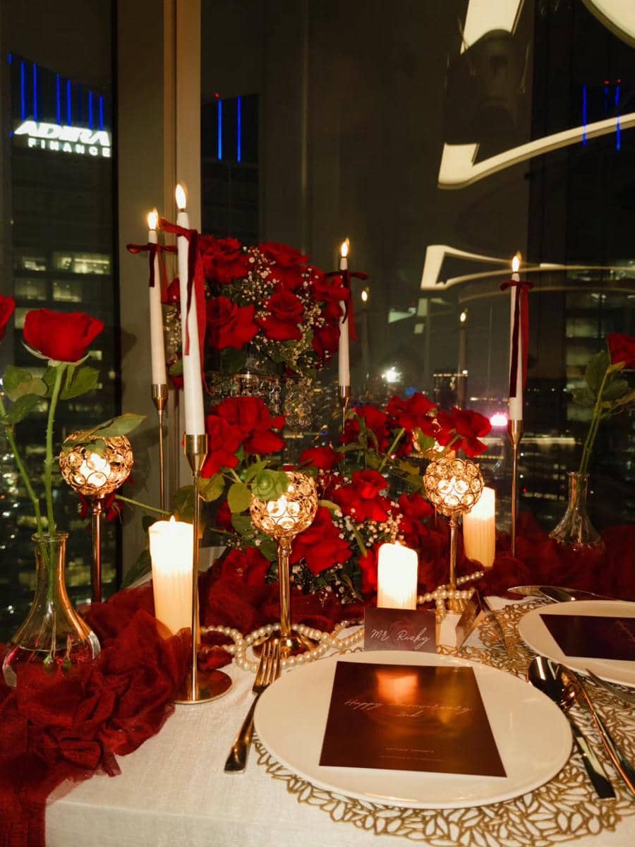 Meja makan romantis dengan lilin menyala, bunga mawar merah, dan dekorasi elegan di restoran dengan pemandangan gedung kota malam hari.