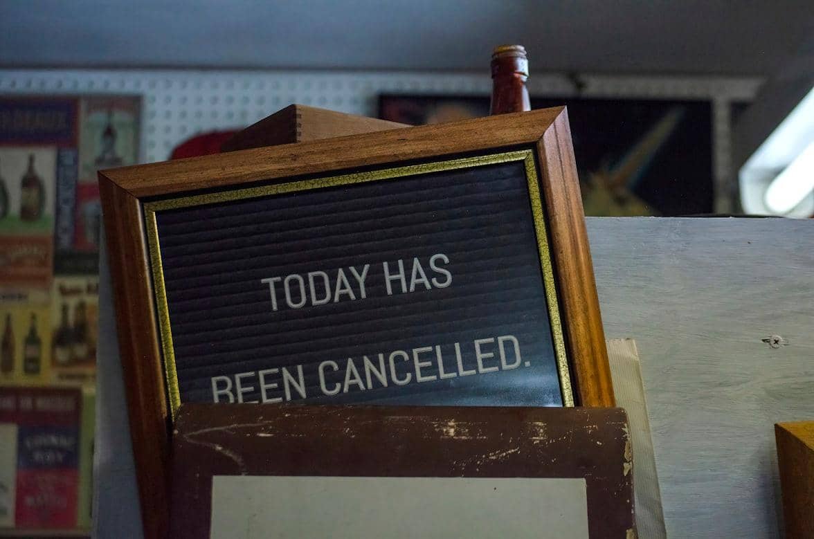 Papan pengumuman berbingkai kayu bertuliskan 'Today Has Been Cancelled' di dalam ruangan dengan latar dinding bergambar poster.