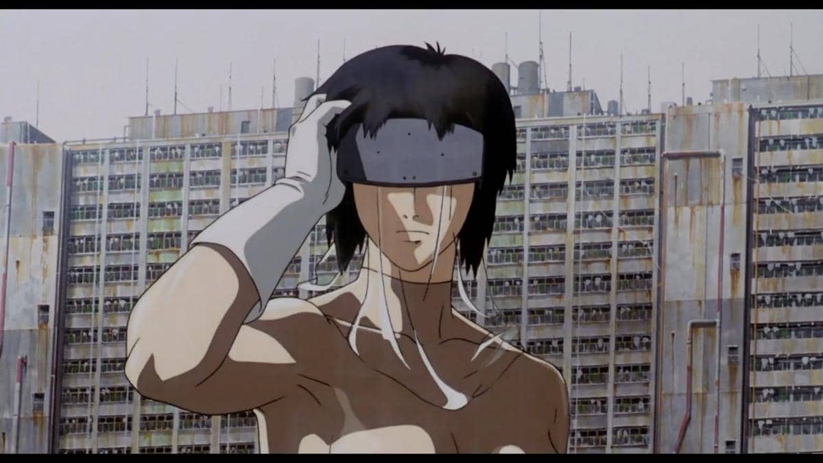 Karakter anime dengan penutup mata logam berdiri di depan gedung tinggi yang tampak usang dalam adegan Ghost in the Shell (1995).