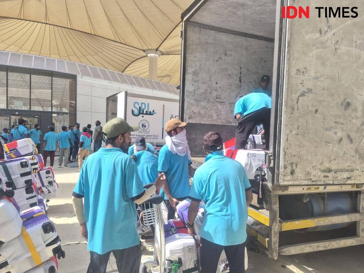 Petugas mengenakan seragam biru sedang memuat koper milik jemaah haji Indonesia ke dalam truk di area logistik.