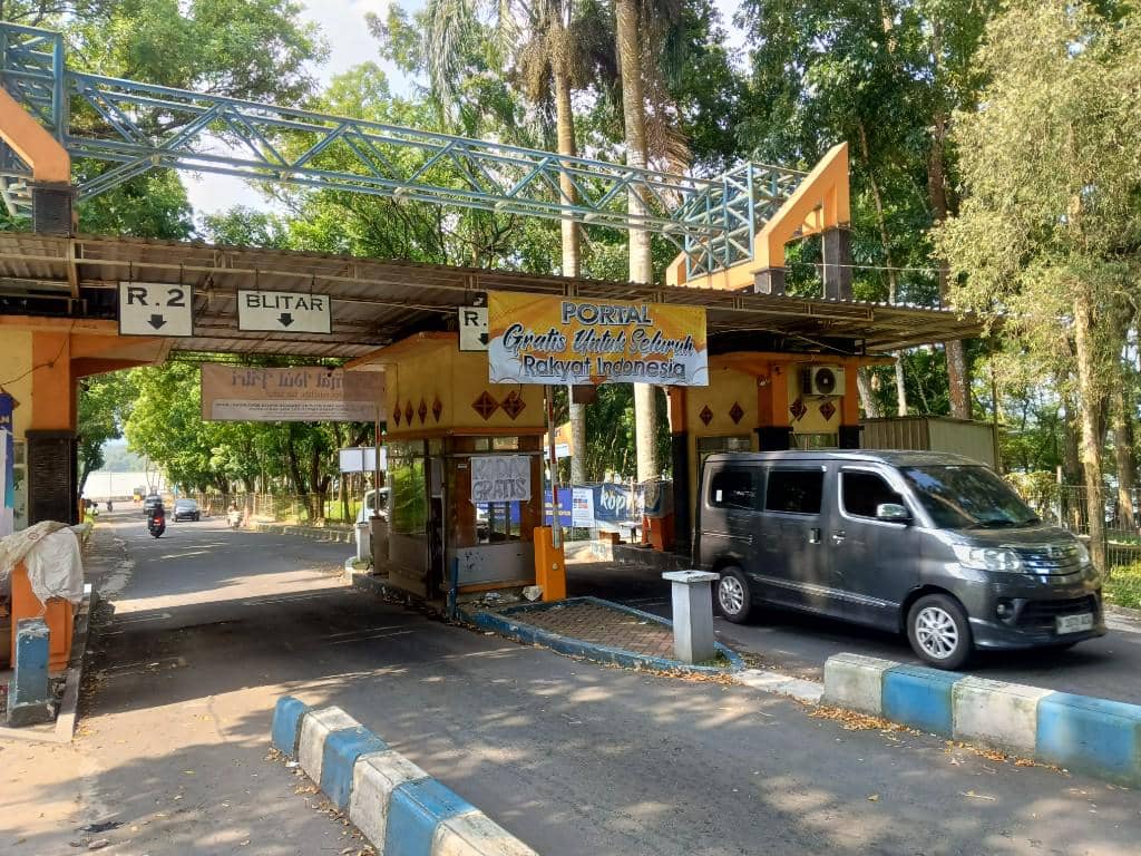 Gerbang masuk Bendungan Lahor di Malang dengan kendaraan melintas dan pepohonan rindang di sekitarnya pada siang hari.