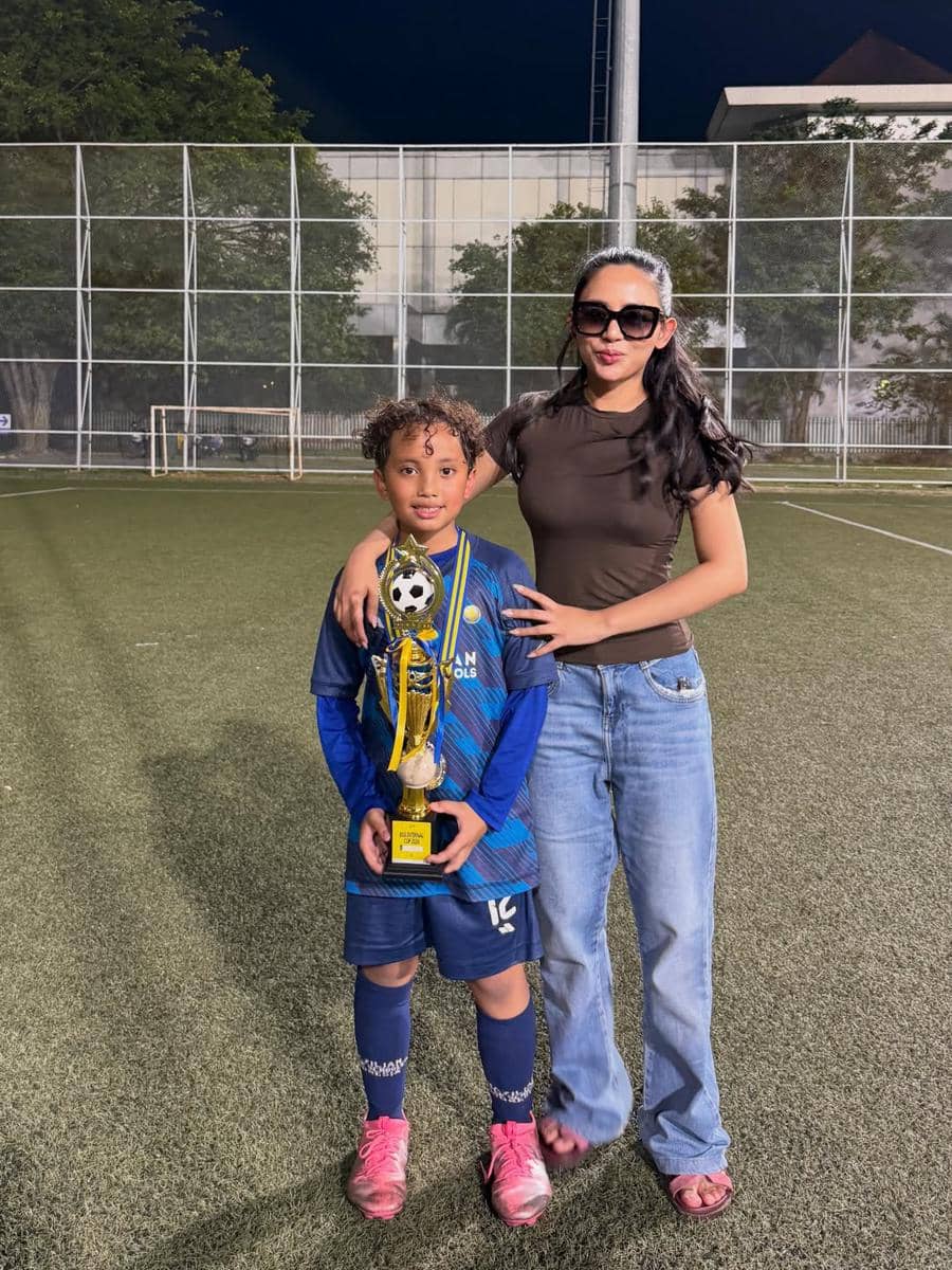 Seorang anak mengenakan seragam sepak bola biru memegang piala sambil berdiri di lapangan bersama seorang wanita berkaus cokelat dan berkacamata hitam.