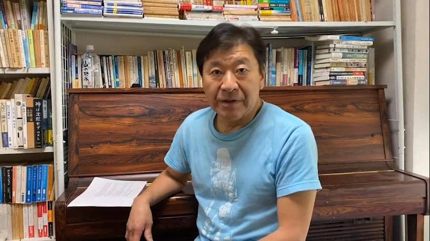 Seorang pria mengenakan kaus biru duduk di depan piano kayu dengan rak buku penuh di belakangnya, tampak berbicara ke arah kamera.