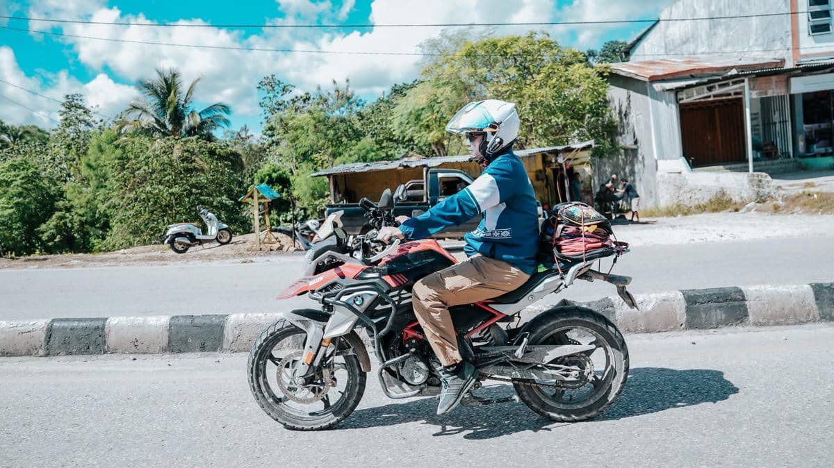 Seorang pengendara motor touring mengenakan jaket biru dan helm putih melintasi jalan dengan latar pepohonan dan bangunan sederhana.