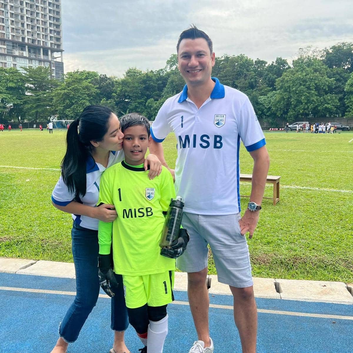 Seorang anak mengenakan seragam sepak bola berwarna hijau bersama dua orang dewasa di tepi lapangan hijau terbuka.