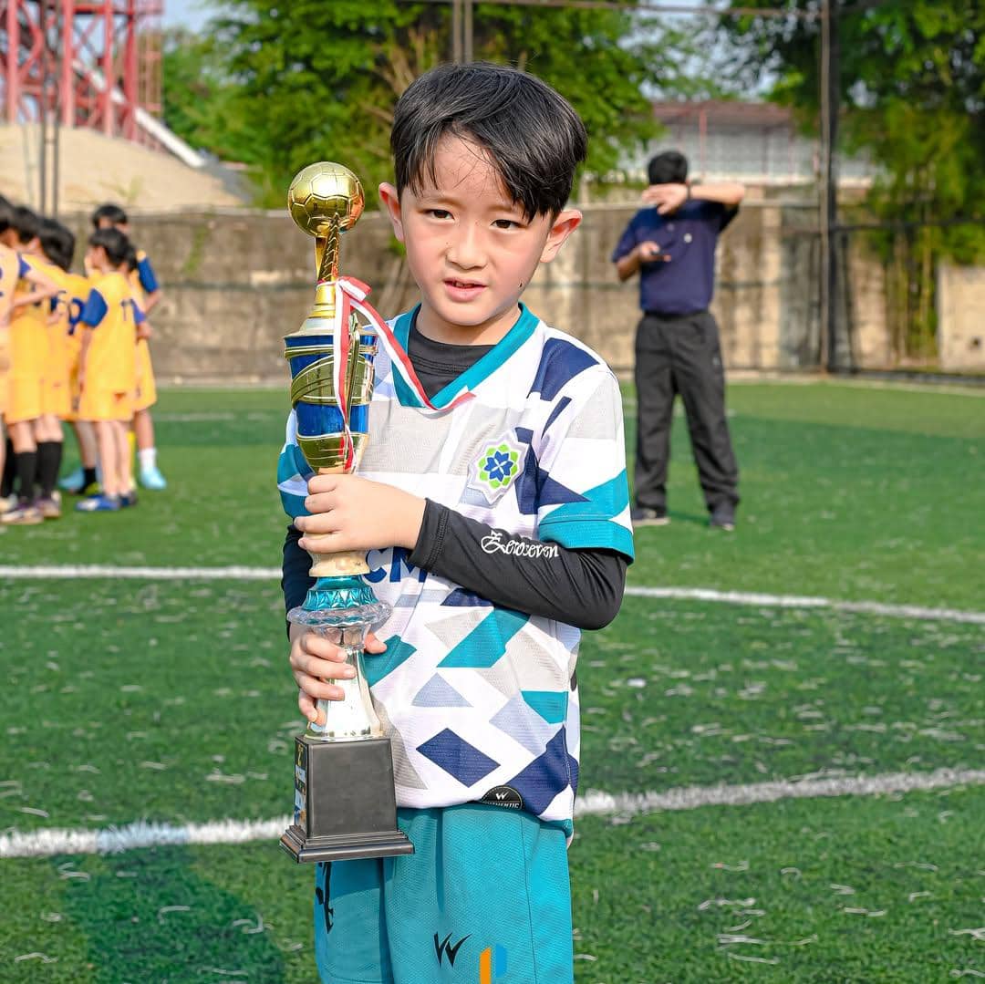 Seorang anak laki-laki mengenakan seragam sepak bola memegang piala di lapangan hijau setelah pertandingan bersama timnya.