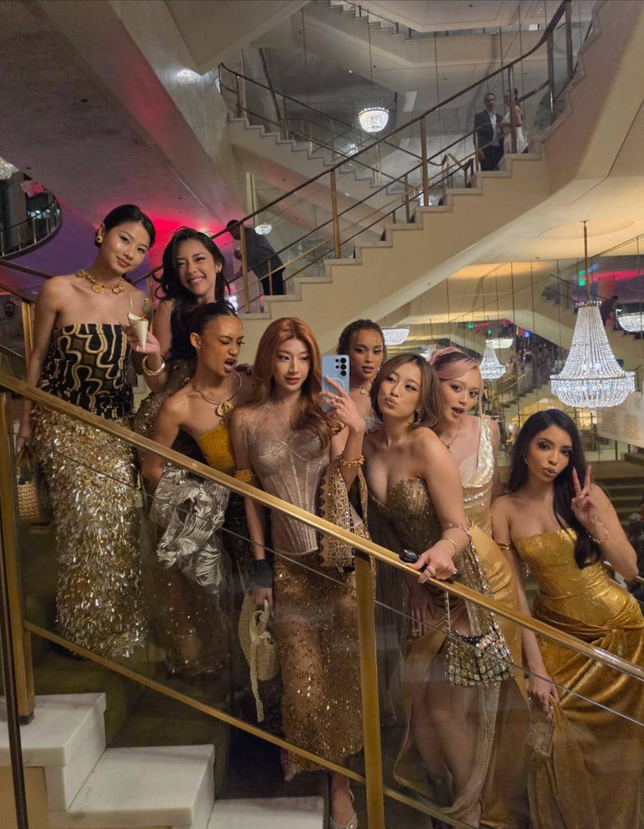 Sekelompok wanita berbusana glamor warna emas berpose di tangga mewah dengan lampu gantung besar di acara Gold House Gold Gala 2026.