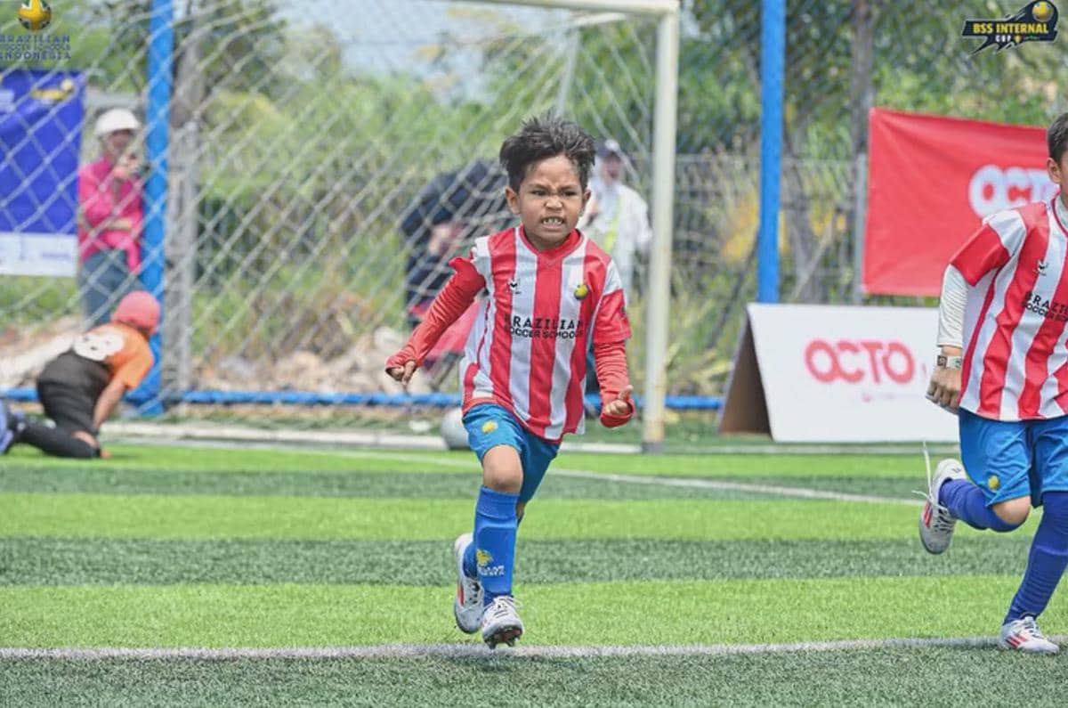 Seorang anak mengenakan seragam sepak bola merah putih berlari di lapangan rumput sintetis dengan ekspresi semangat saat pertandingan.