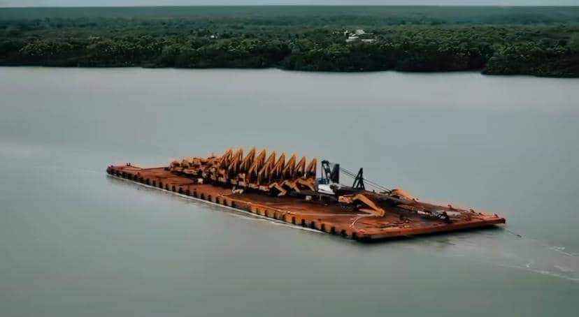 Sebuah tongkang besar membawa peralatan konstruksi berwarna oranye melintasi perairan luas dengan latar hutan hijau di kejauhan.