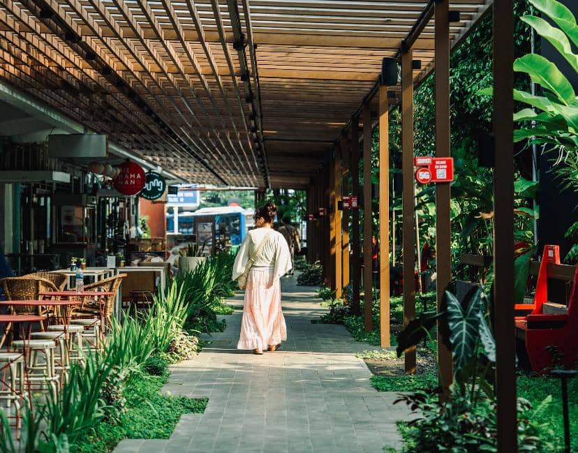 Seorang perempuan berjalan di koridor terbuka M Bloc Group yang dikelilingi tanaman hijau dan area duduk di sisi kiri dan kanan.