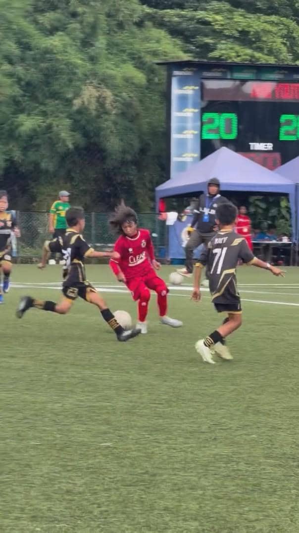 Seorang anak mengenakan seragam merah bermain sepak bola di lapangan hijau melawan tim berbaju hitam dengan skor pertandingan terlihat di latar belakang.