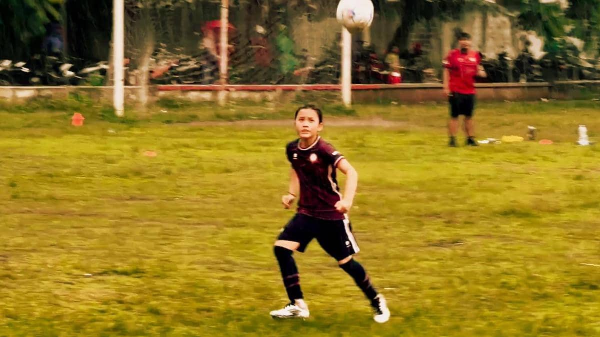 Seorang anak mengenakan seragam sepak bola berwarna merah tua sedang berlari mengejar bola di lapangan rumput terbuka.
