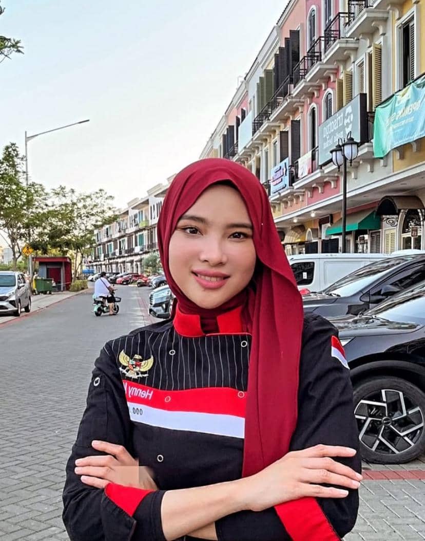Seorang perempuan berhijab merah mengenakan seragam koki hitam-merah dengan tangan terlipat berdiri di jalan kota dengan deretan ruko warna-warni.