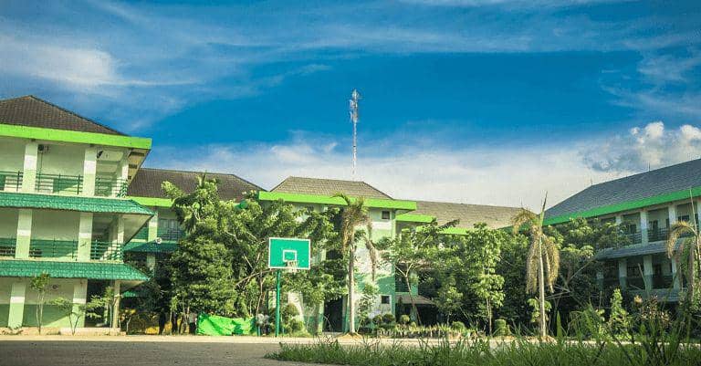 Gedung sekolah bertingkat dengan cat hijau dan putih, dikelilingi pepohonan serta lapangan basket di bawah langit biru cerah.