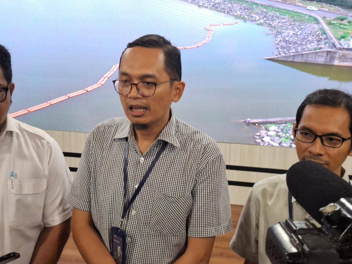 Seorang pria berbicara di depan mikrofon bersama dua orang lainnya di dalam ruangan dengan latar belakang gambar waduk besar.