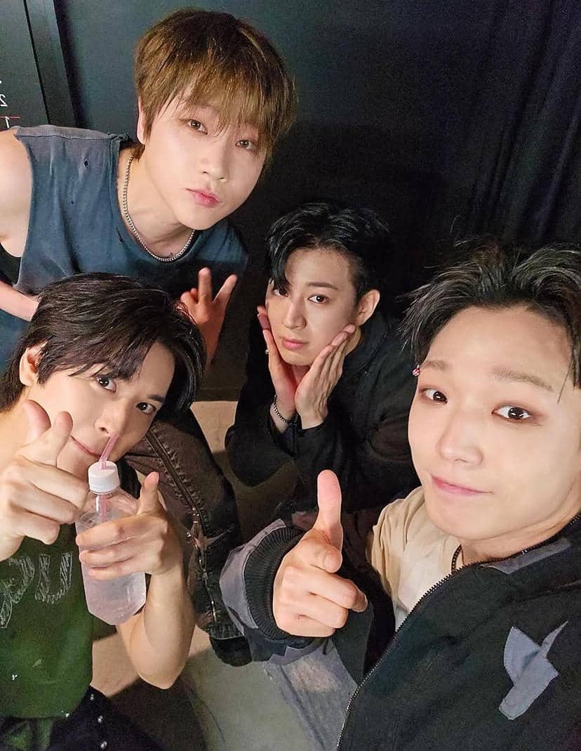 Empat anggota grup musik iKON berpose santai bersama di belakang panggung dengan ekspresi ceria dan gaya kasual.
