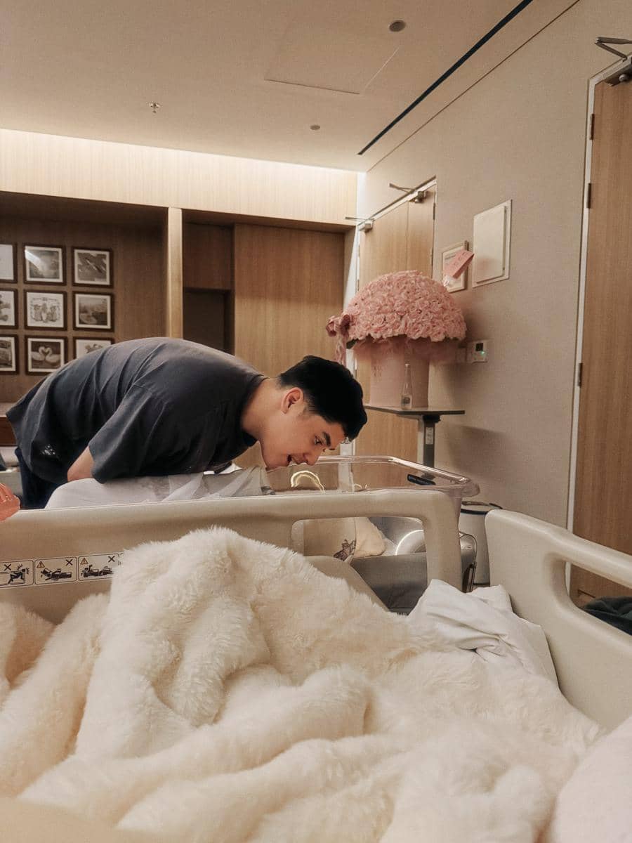 Seorang pria membungkuk melihat bayi yang terbaring di boks bayi di kamar rumah sakit dengan dekorasi bunga berwarna merah muda.