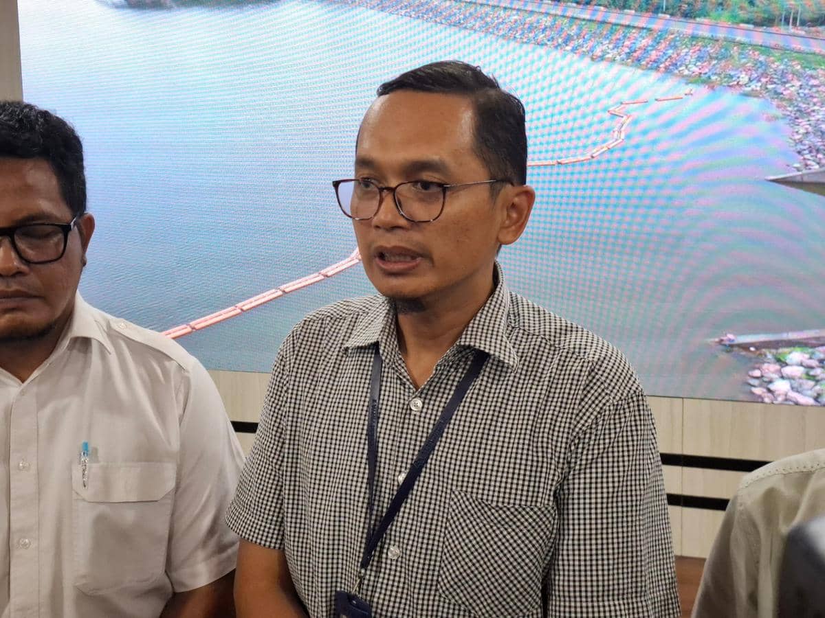 Seorang pria berkemeja kotak-kotak berbicara di depan layar besar yang menampilkan pemandangan waduk atau bendungan.