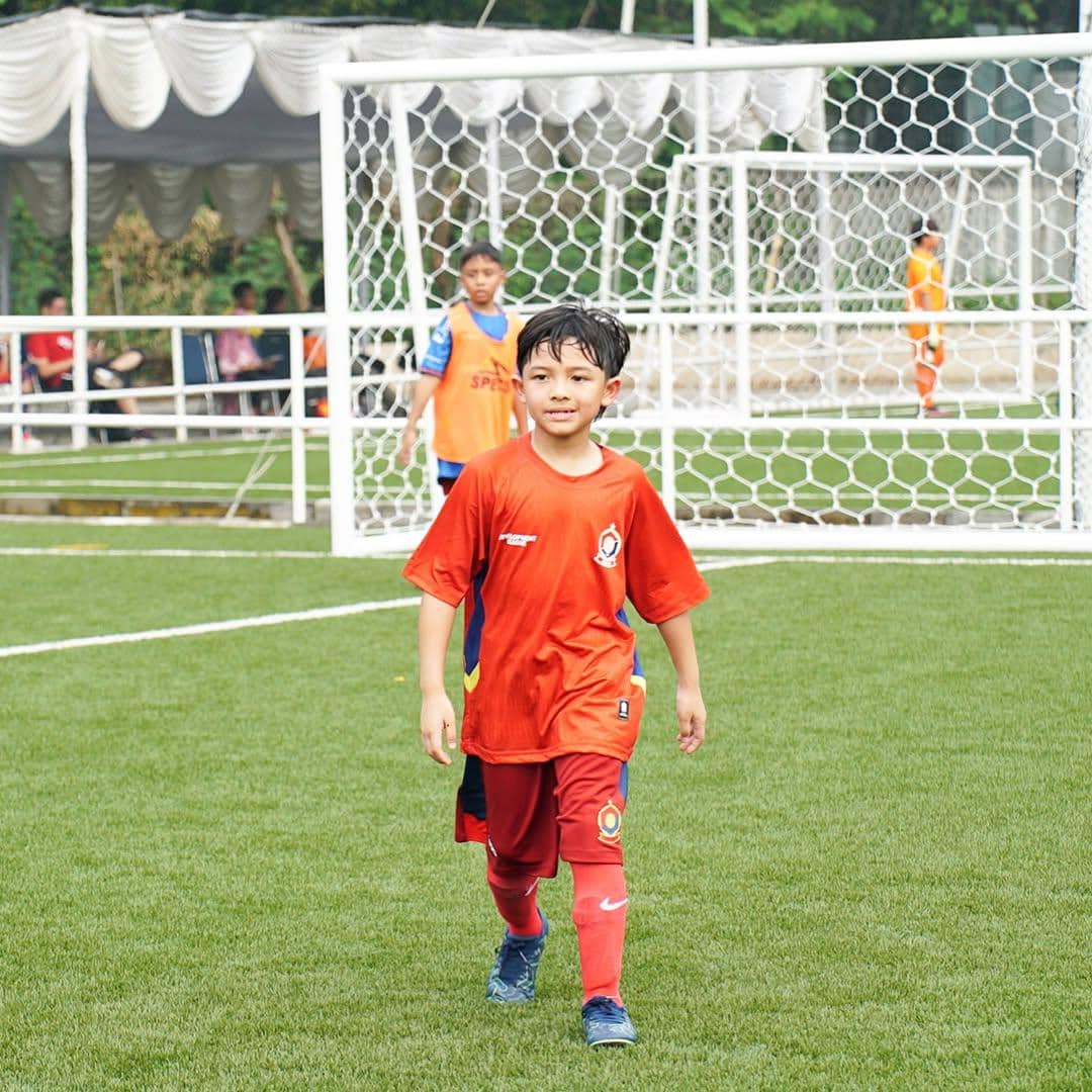 Seorang anak laki-laki mengenakan seragam sepak bola merah berjalan di lapangan rumput dengan gawang di latar belakang.