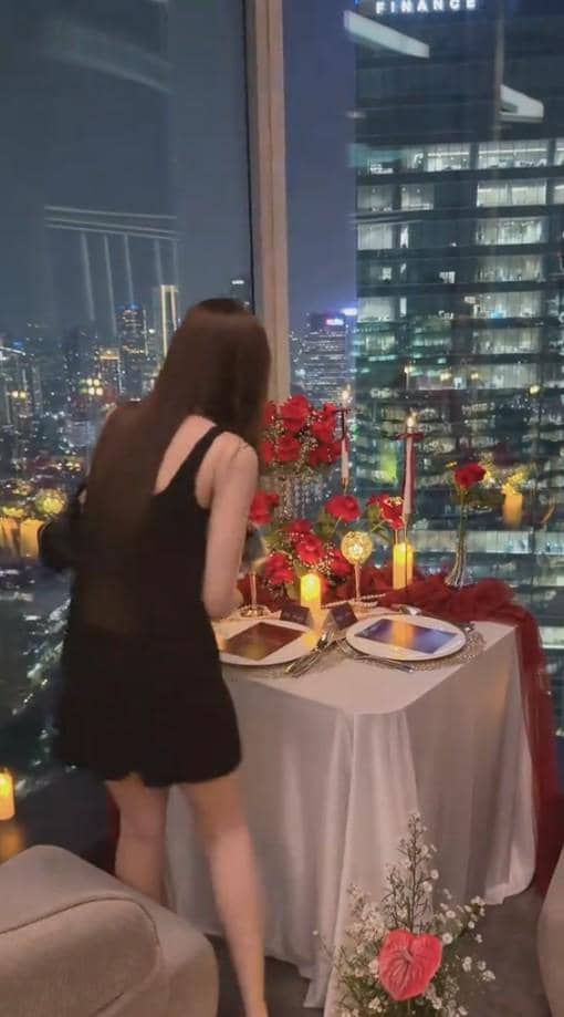 Seorang wanita mengenakan gaun hitam berdiri di depan meja makan romantis dengan dekorasi bunga mawar merah dan lilin di ruangan tinggi berlatar gedung kota malam hari.
