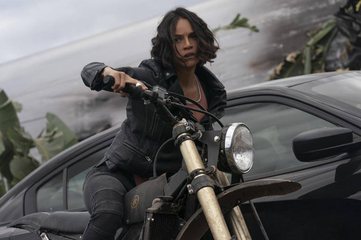 Seorang perempuan mengenakan jaket kulit hitam mengendarai motor trail di samping mobil hitam dalam adegan film F9: The Fast Saga.
