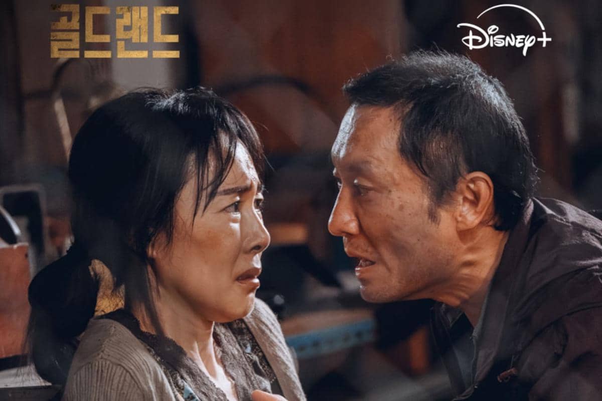 Dua karakter dalam adegan tegang dari drama Korea Gold Land, menatap satu sama lain dengan ekspresi emosional di ruangan redup.