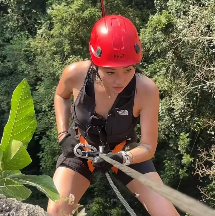Seorang perempuan mengenakan helm merah dan perlengkapan panjat tebing sedang menuruni tebing dengan tali di area hijau Gua Ngeleng.