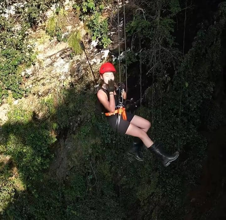 Seorang perempuan mengenakan helm merah dan perlengkapan keselamatan sedang bergelantungan di tebing gua yang dipenuhi tanaman hijau.