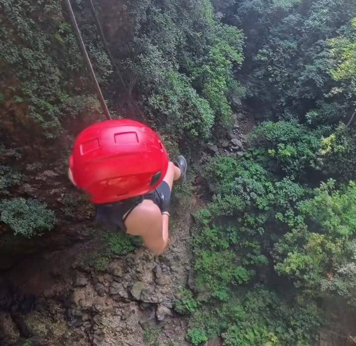 Seorang perempuan mengenakan helm merah sedang menuruni tebing curam di Gua Ngeleng yang dikelilingi pepohonan hijau lebat.
