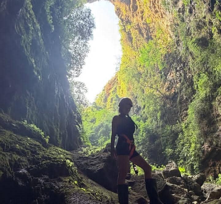 Seorang perempuan berdiri di dalam Gua Ngeleng yang tinggi dengan dinding batu curam dan cahaya alami masuk dari lubang di atasnya.
