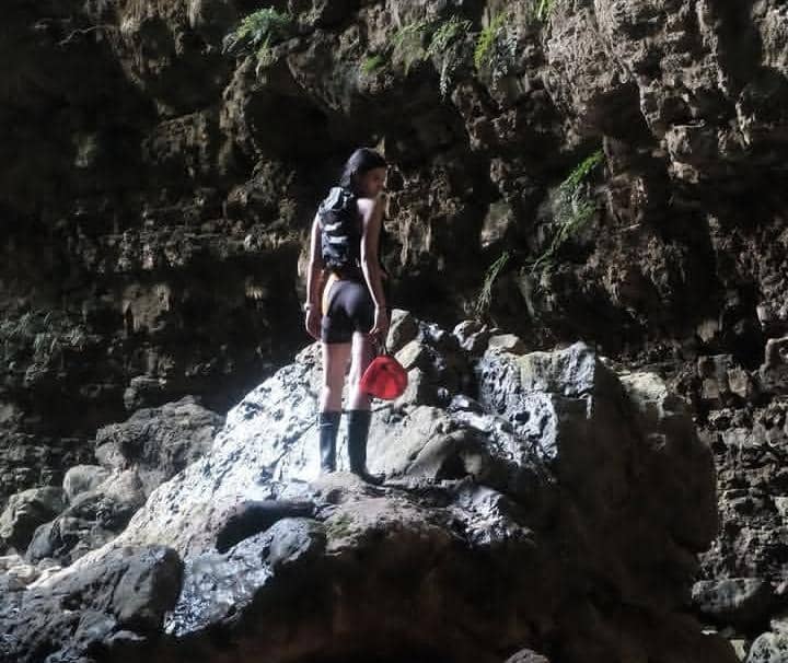 Seorang perempuan berdiri di atas batu besar di dalam gua dengan dinding batu alami, mengenakan ransel dan sepatu bot hitam.