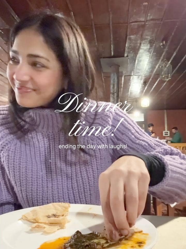 Seorang wanita mengenakan sweter ungu tersenyum sambil menikmati makan malam di restoran dengan suasana hangat dan pencahayaan lembut.