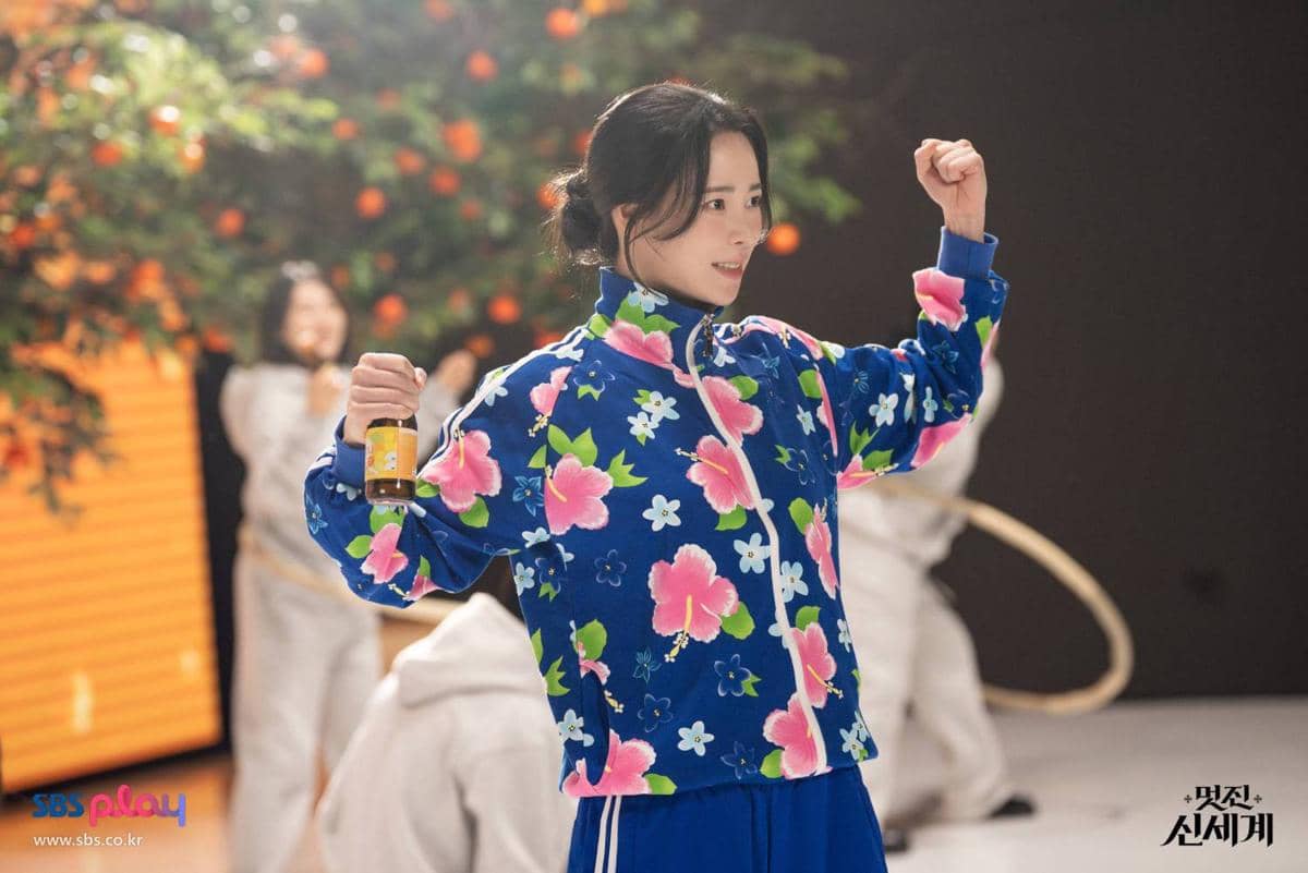 Seorang wanita mengenakan jaket biru bermotif bunga berpose dengan tangan terangkat sambil memegang botol di lokasi syuting drama Korea My Royal Nemesis.