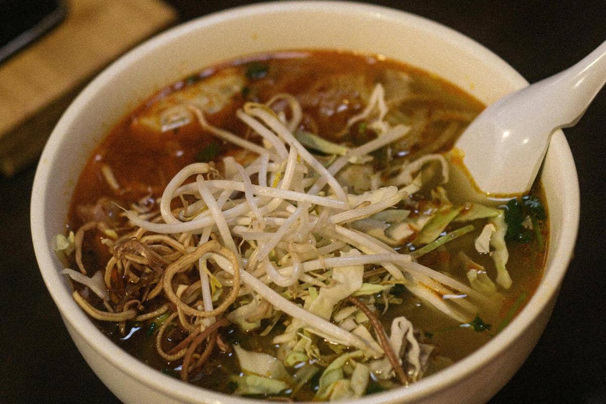 Ilustrasi semangkuk lontong mie khas Surabaya dengan kuah petis dan taburan ebi di area food court pasar tradisional.