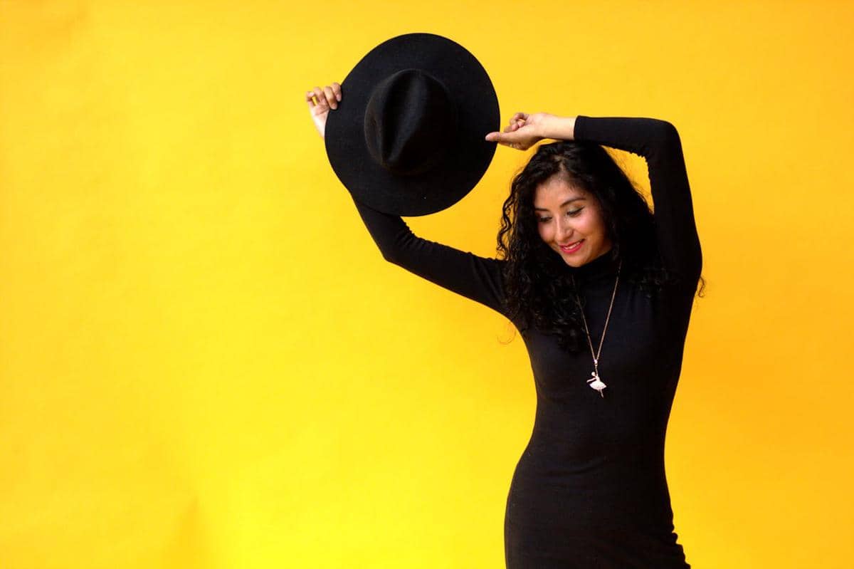 Seorang wanita mengenakan dress hitam lengan panjang model turtle neck sambil memegang topi hitam di depan latar belakang kuning cerah.