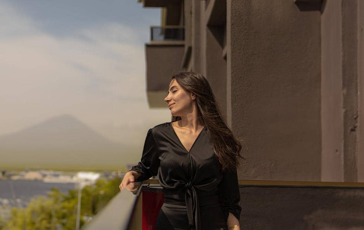 Seorang wanita mengenakan dress hitam berpotongan v-neck berdiri di balkon dengan latar bangunan dan pemandangan pegunungan di kejauhan.