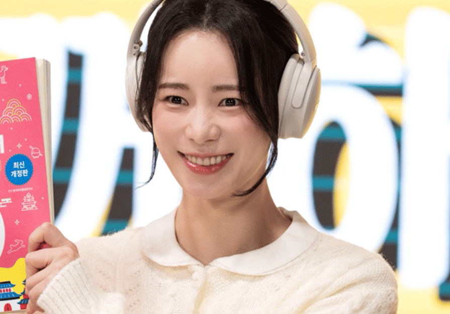 Seorang wanita mengenakan headphone putih sambil tersenyum dan memegang buku berwarna merah muda dalam cuplikan drama Korea My Royal Nemesis.