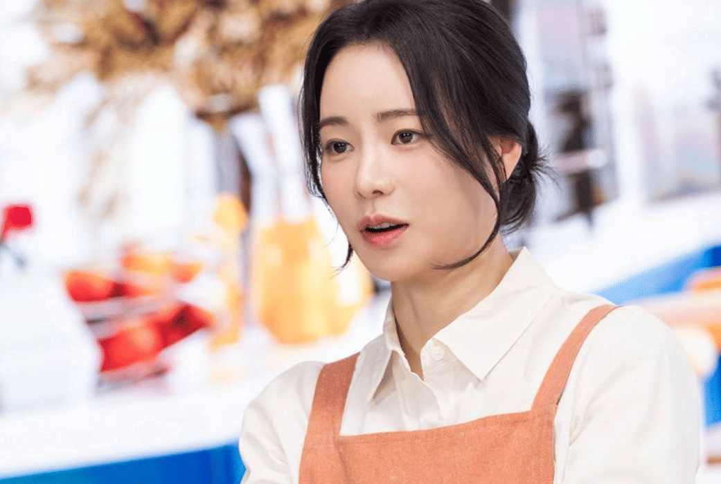 Seorang wanita mengenakan kemeja putih dan apron oranye tampak berbicara di latar ruangan dengan warna cerah dalam cuplikan drama Korea My Royal Nemesis.