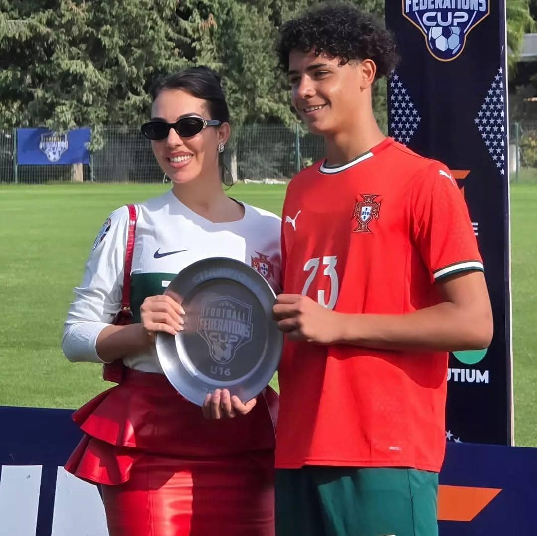 Seorang wanita mengenakan jersey Portugal berwarna putih berpose bersama pemain muda yang memakai jersey merah Portugal sambil memegang trofi di lapangan sepak bola.