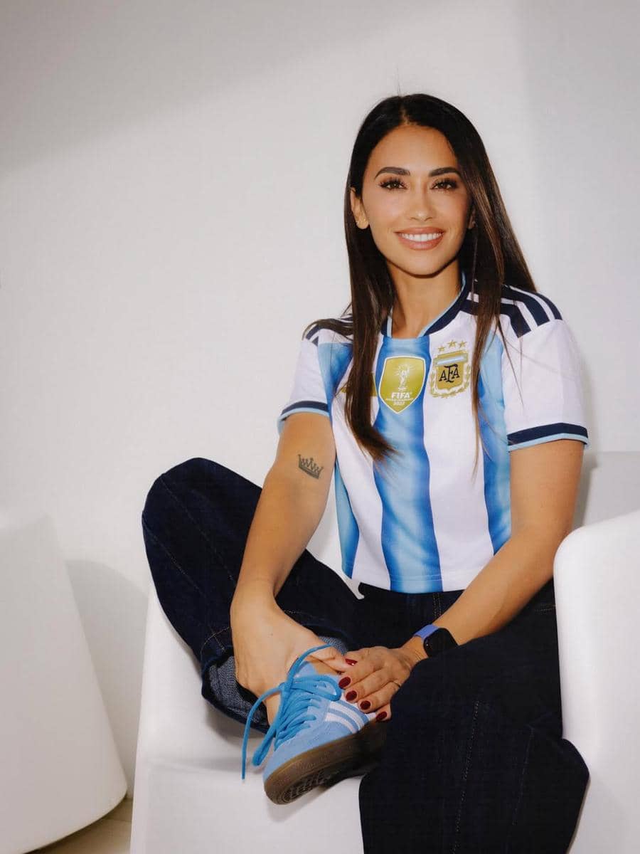 Seorang wanita mengenakan jersey tim nasional Argentina berwarna biru putih dengan celana jeans gelap dan sepatu biru, duduk santai di kursi putih.