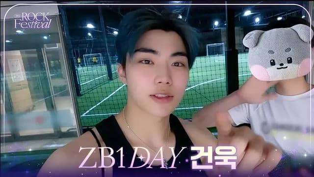 Seorang pria berambut gelap mengenakan tank top hitam berpose di depan lapangan futsal dalam ruangan bersama seseorang yang wajahnya tertutup boneka.