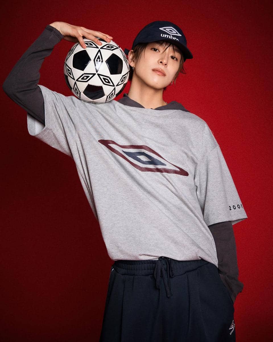 Seorang model mengenakan topi dan kaus abu-abu bermotif logo Umbro sambil memegang bola sepak di bahu dengan latar belakang merah.