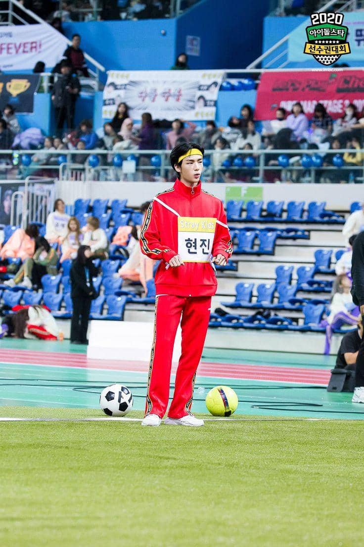 Seorang peserta mengenakan tracksuit merah dengan ikat kepala kuning berdiri di lapangan indoor ISAC 2020 dengan dua bola di depannya.