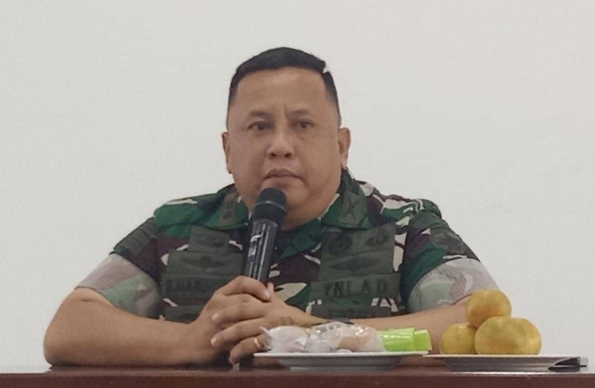 Seorang perwira TNI mengenakan seragam loreng duduk di meja sambil memegang mikrofon dengan beberapa makanan ringan di depannya.