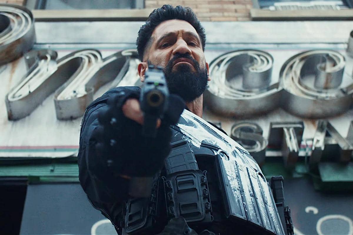 Seorang pria berjanggut mengenakan rompi pelindung hitam mengarahkan pistol ke depan dalam adegan The Punisher: One Last Kill.